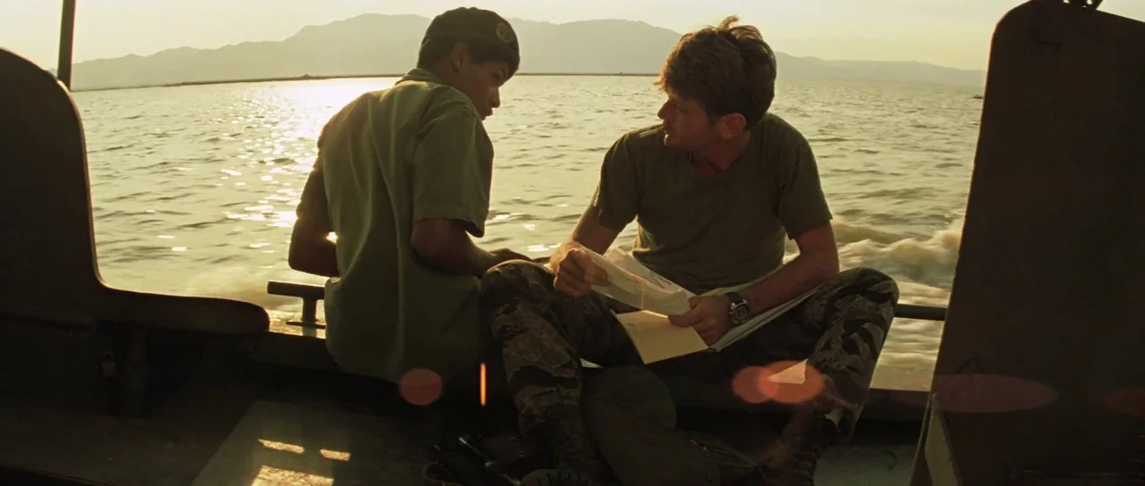 Apocalypse Now (1979) movie still: medium — Two men sit on a small boat moving across a wide lake during sunset. One man reads a letter while the…