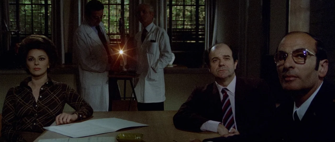 What Have They Done To Your Daughters? (1974) movie still: medium — A dim conference room has three people at a table and two people standing. A…