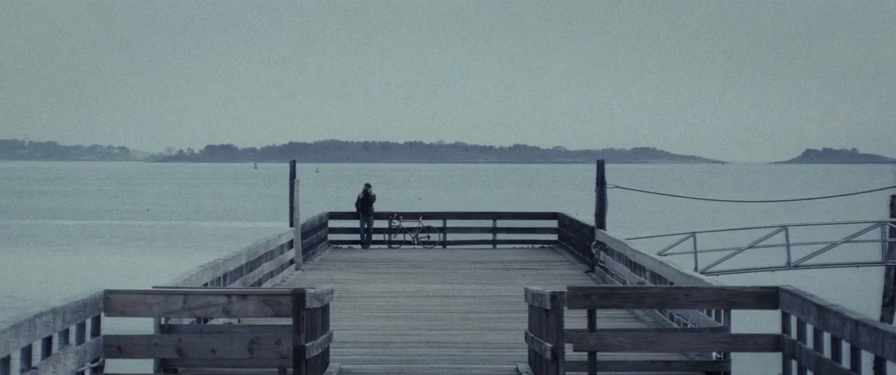 Lords Of Salem (2013) movie still: wide — A long wooden pier extends over a wide, grey expanse of water toward distant land. A lone person stands at…