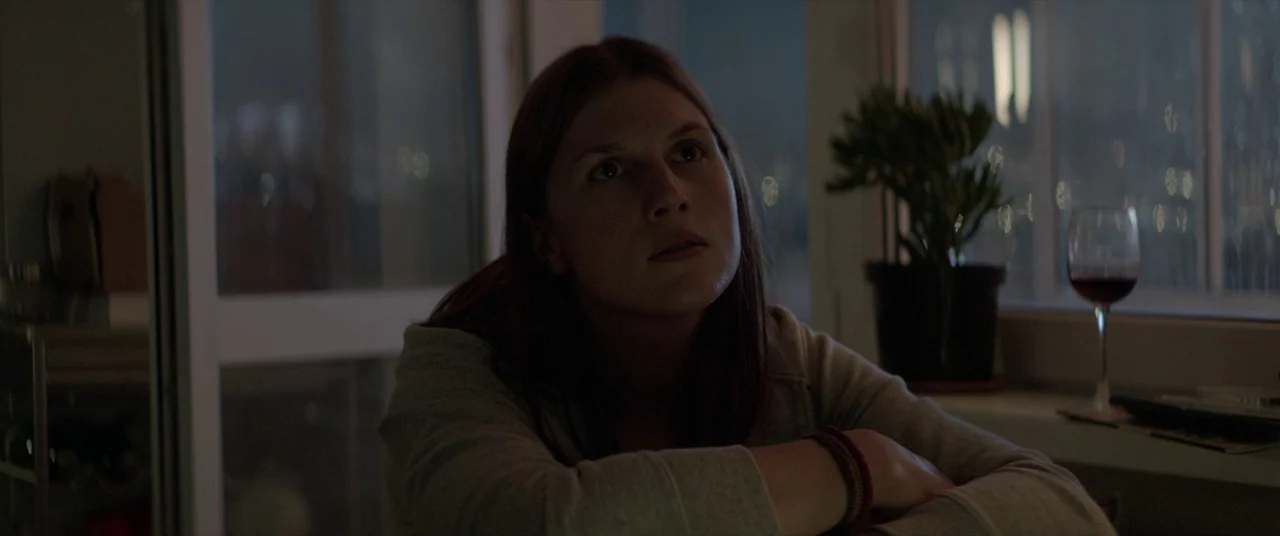Loveless (2017) movie still: medium — An apartment interior at night with a large window facing city lights. A young woman sits by the window…