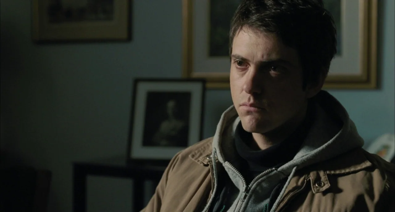 Incendies movie still: medium — A dimly lit room with framed pictures hanging on blue-grey walls. A man wearing a layered jacket sits and stares…