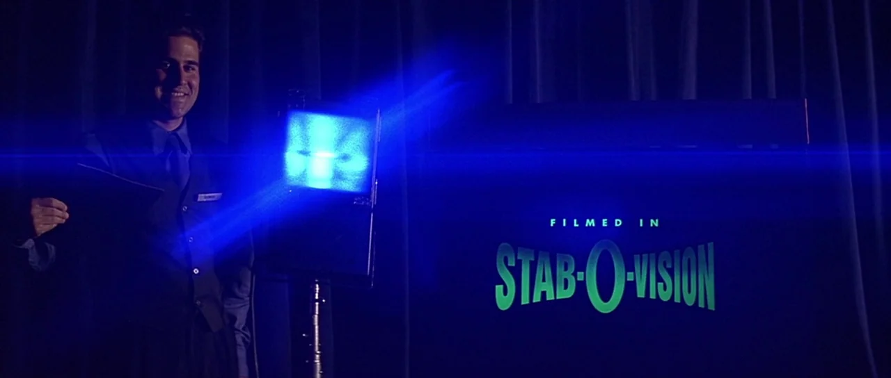 Scream 2 (1997) movie still: medium — A man in a vest stands on a dark stage with heavy fabric curtains. A glowing screen next to him displays green…