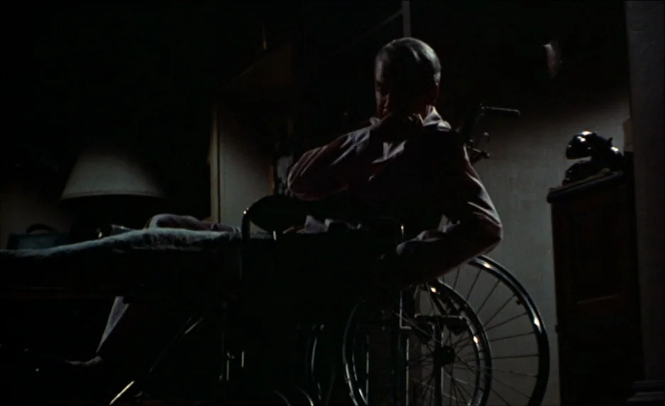 Rear Window (1954) movie still: medium — A dark study contains a wheelchair with a man sitting in the center. The man rests one arm on the chair…