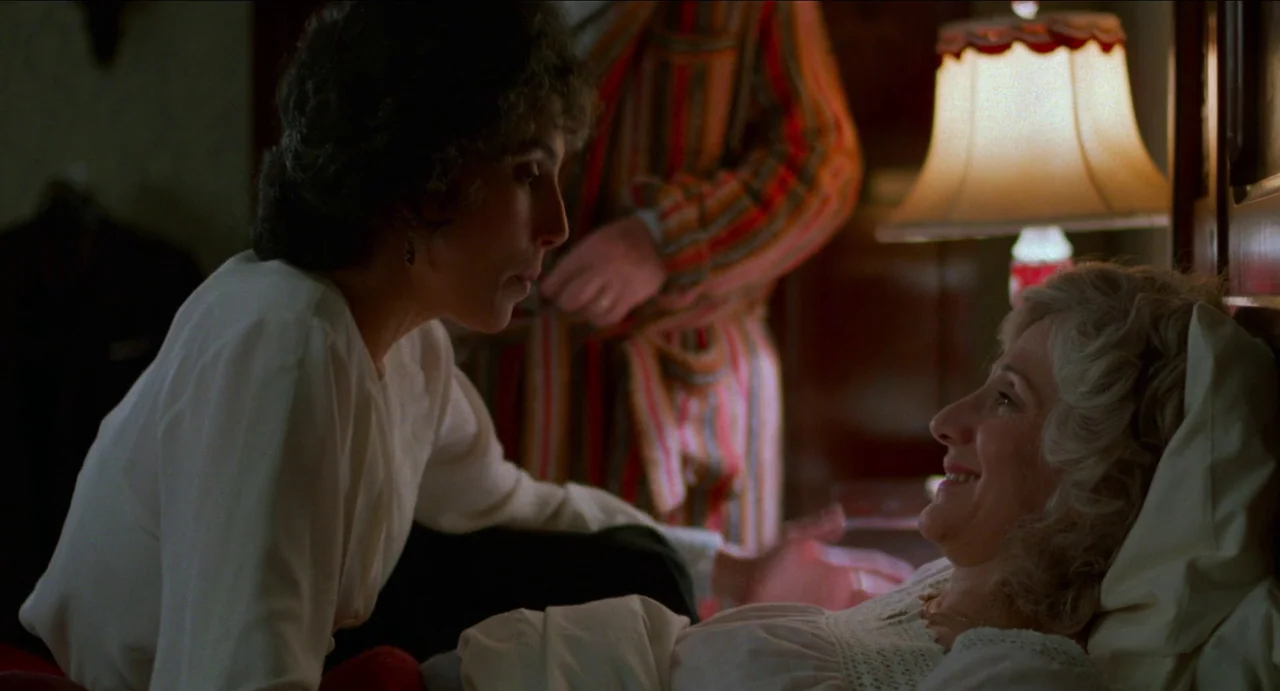 Moonstruck (1987) movie still: medium — A dimly lit room with a bedside lamp shining over two women talking. One woman leans over the bed while the…
