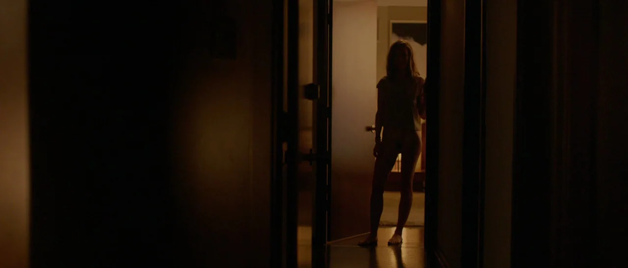 The Invitation (2015) movie still: fullbody — A dark hallway with a single open doorway leading into a lit space. A woman stands centered in the…
