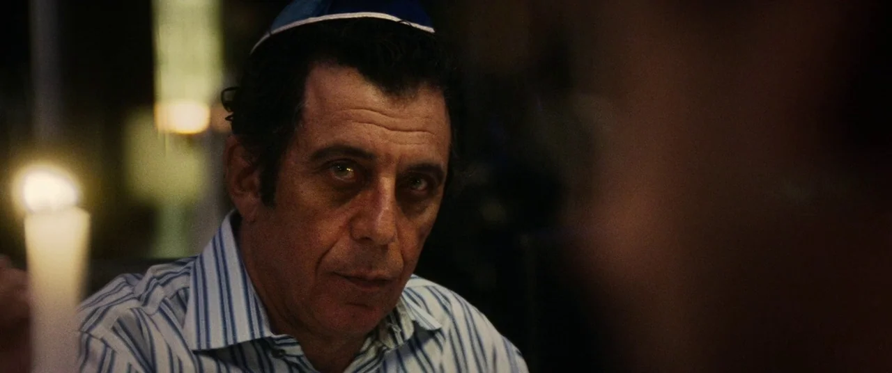 Uncut Gems (2019) movie still: closeup — A man sits in a dimly lit space with a candle in the foreground. He wears a yarmulke and gazes forward…