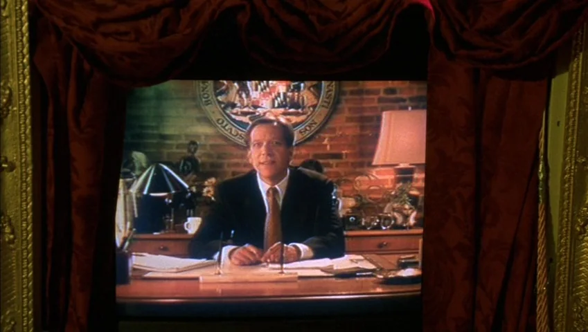 Making of: Castle Freak (1995) movie still: medium — A man in a suit is displayed on a television screen framed by dark curtains. He sits behind a…