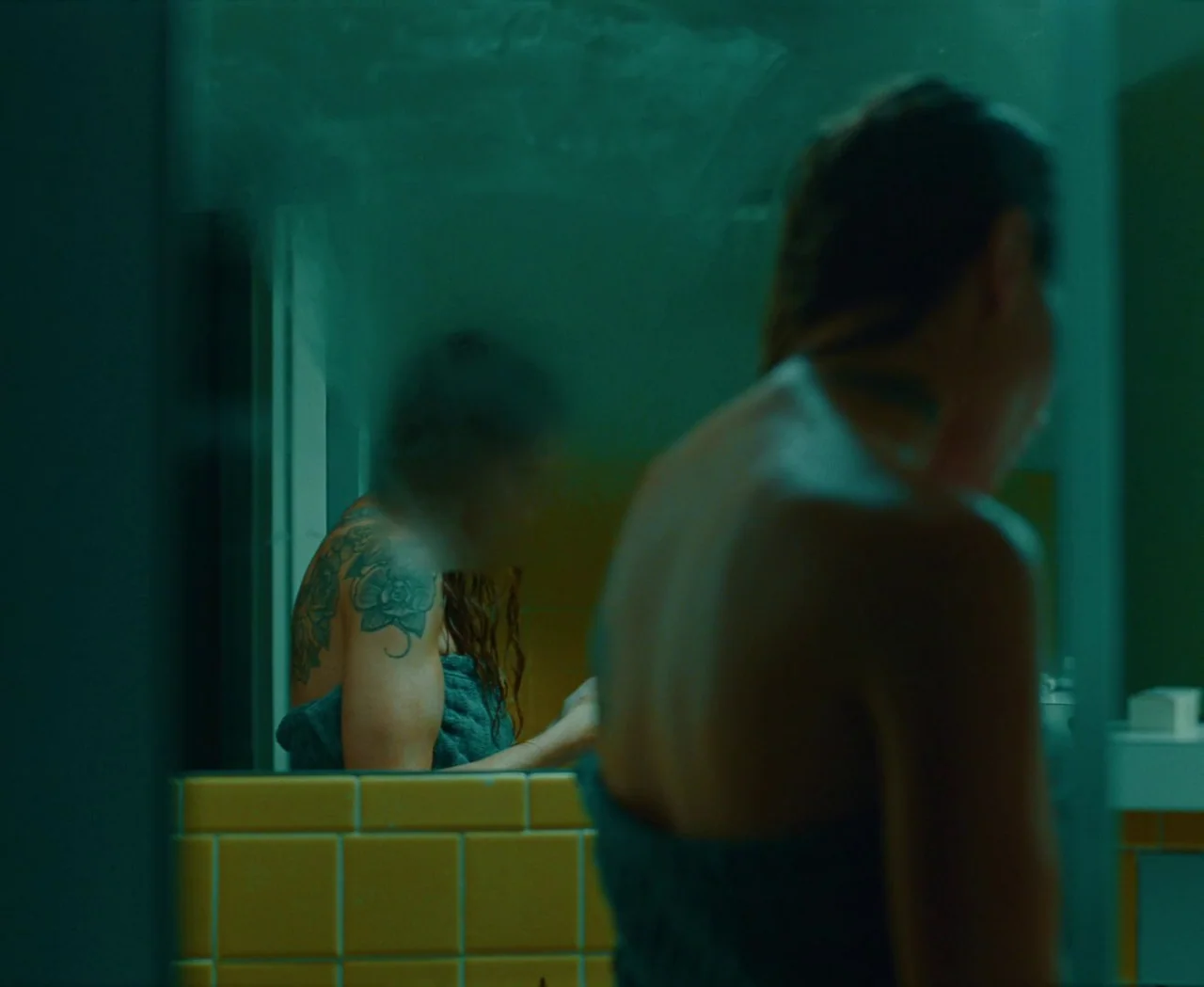 Monica (2022) movie still: medium — A bathroom with yellow square wall tiles is reflected in a mirror. A woman with a shoulder tattoo stands in the…