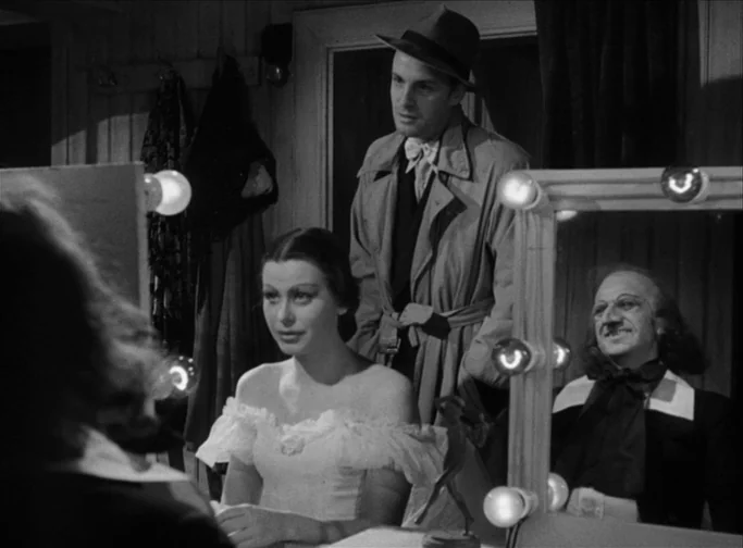 Summer Interlude (1951) movie still: medium — A dark, cramped space lit by vanity lights surrounding a rectangular mirror frame. A woman in a dress…
