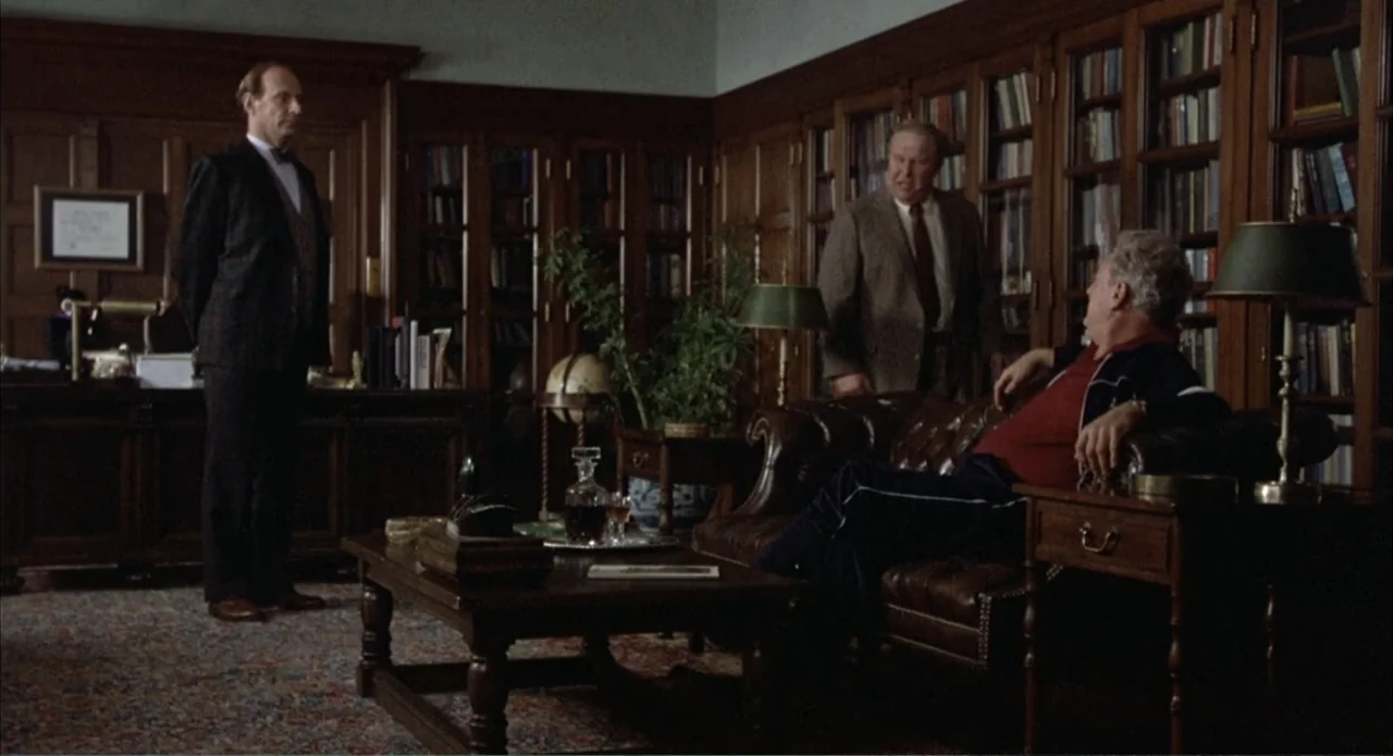 Back to School (1986) movie still: wide — A wood-paneled room features wall-to-wall bookshelves filled with books and a dark leather sofa. Three men…
