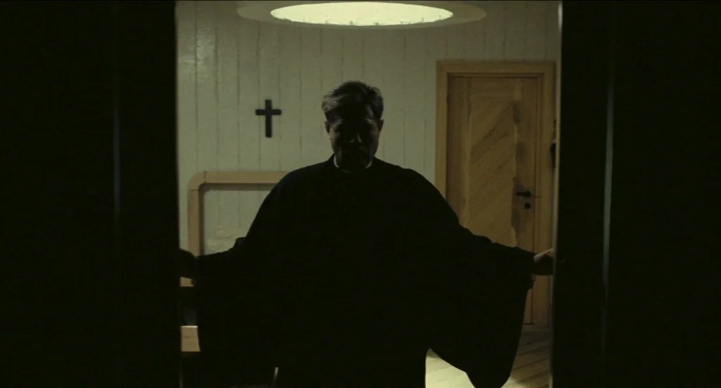 Todo Modo (1976) movie still: medium — A spare, light-colored room features vertical wood-paneled walls and a black cross mounted centrally. A man…