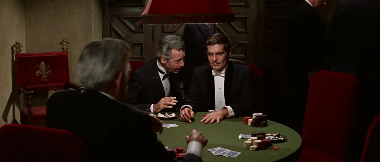 Funny Girl (1968) movie still: medium — A dimly lit room features a round green felt table surrounded by red chairs. Three men in formal tuxedos are…