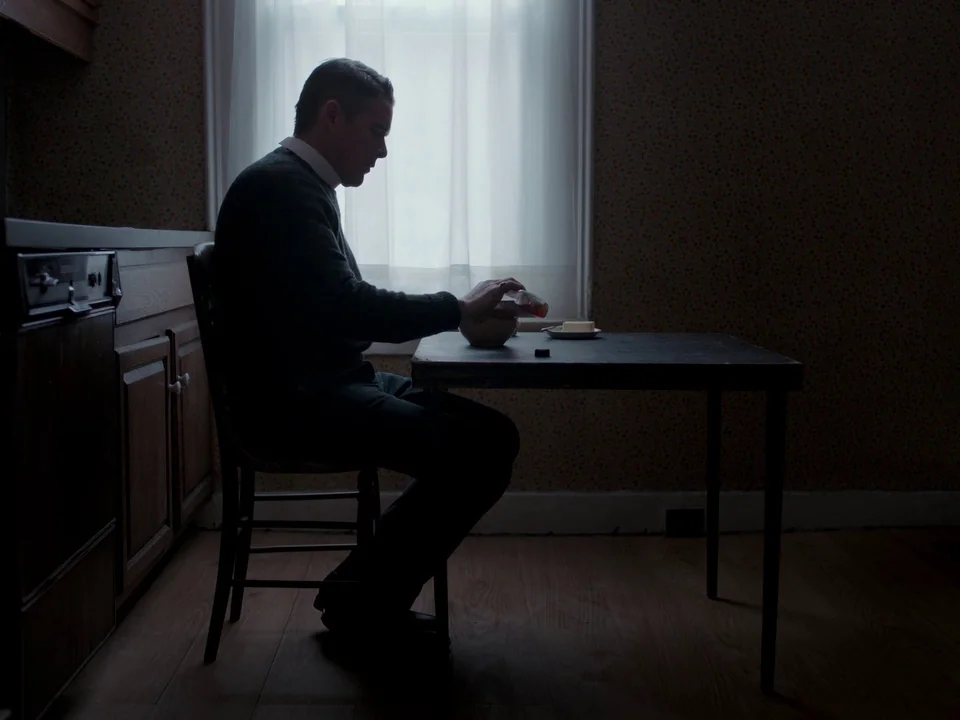 First Reformed (2017) movie still: medium — A dark dining room with patterned wallpaper features a man sitting at a table. He pours liquid into a…