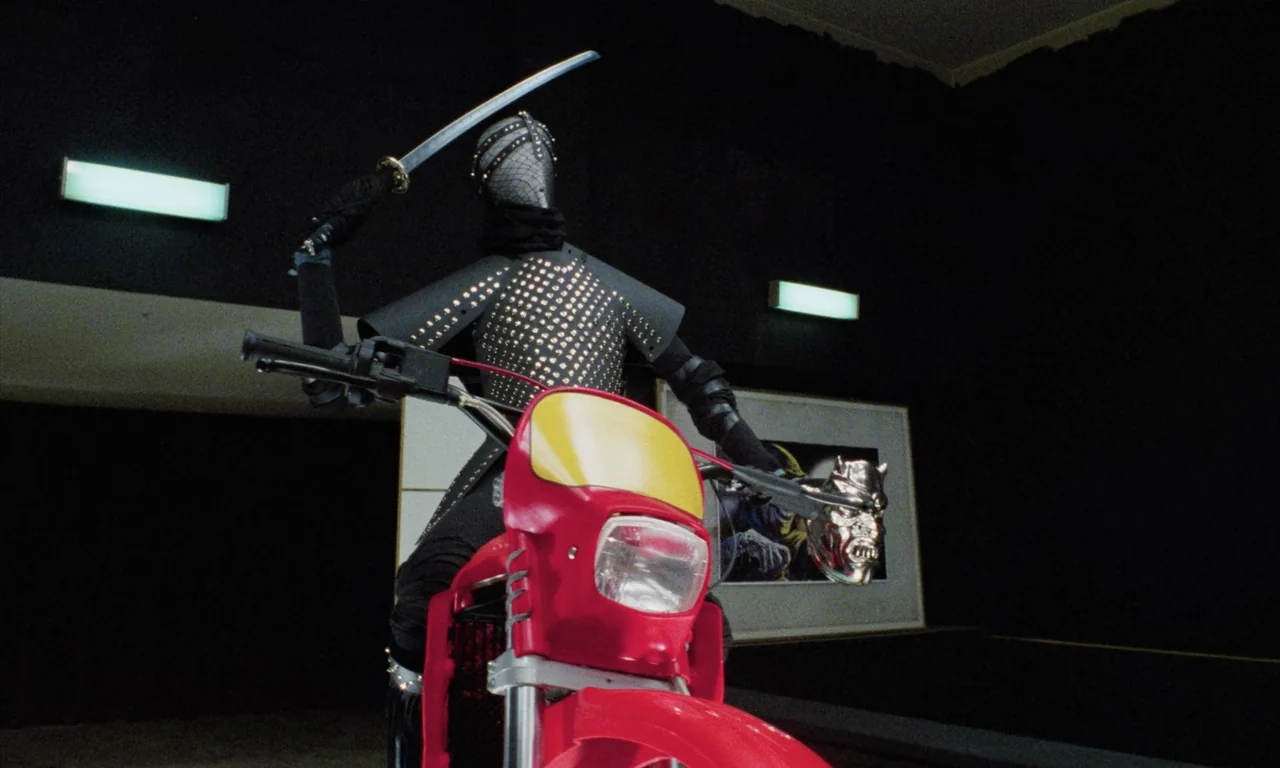 Demons (1985) movie still: fullbody — A dark exhibition space with black walls and rectangular light fixtures. A figure in studded armor sits on a…