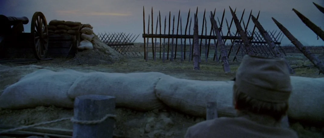 Cold Mountain (2003) movie still: medium — A field with sharpened wooden stakes and sandbag earthworks under a grey morning sky. A soldier from…