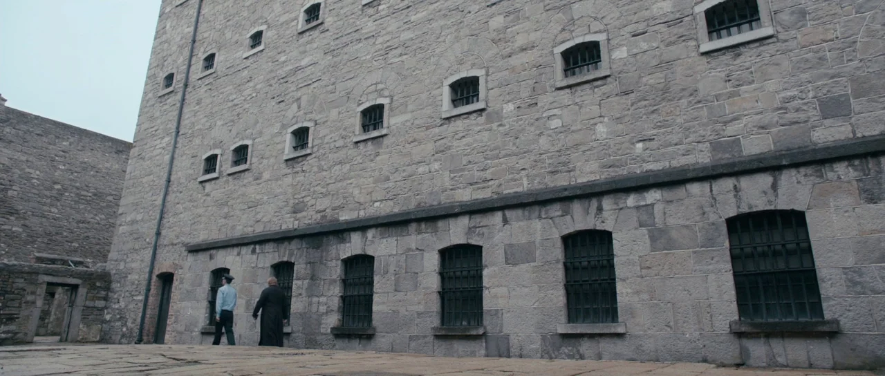 Calvary (2014) movie still: wide — An imposing stone structure with multiple barred windows stands over a cobblestone area. A uniformed guard and a…