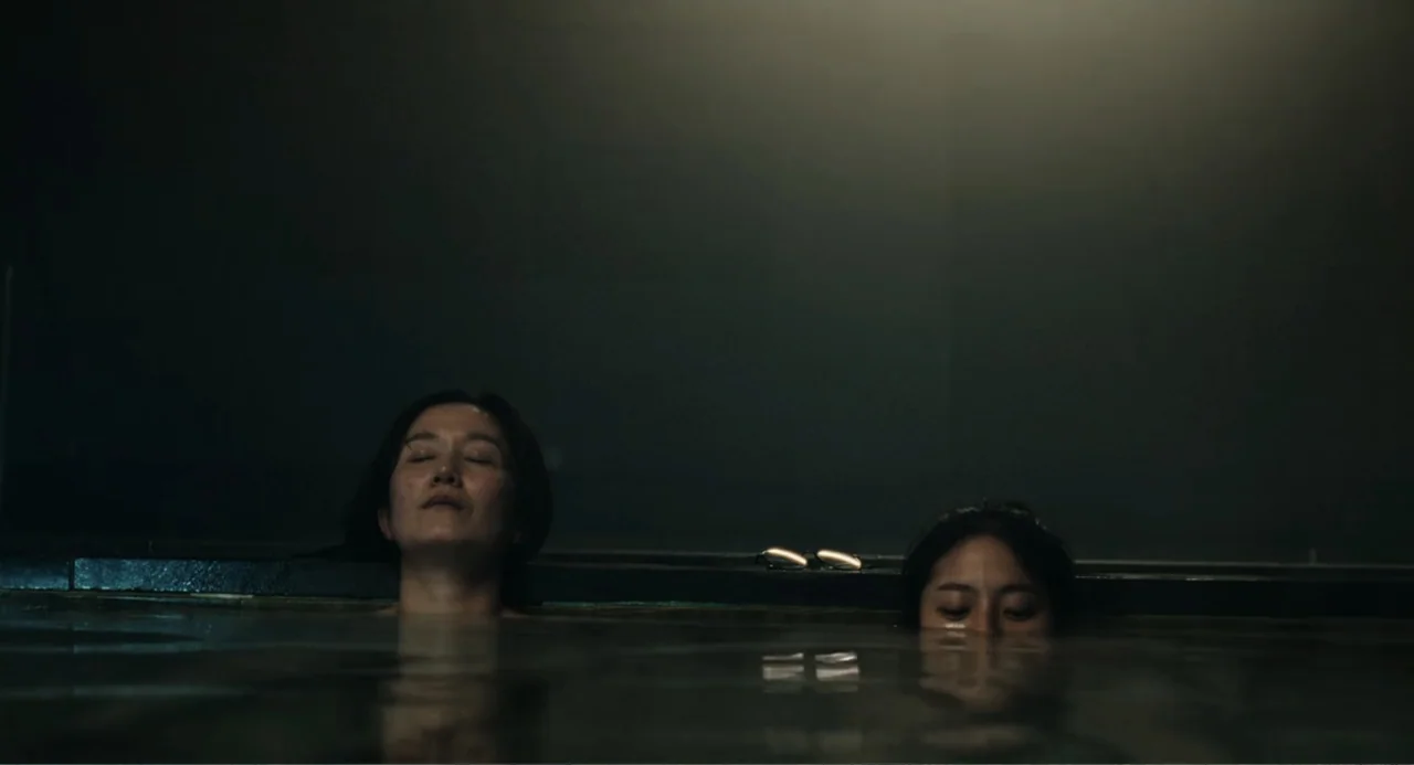 Winter in Sokcho (2025) movie still: medium — Two women are submerged in a dark stone pool of water at night. They rest with their heads visible…