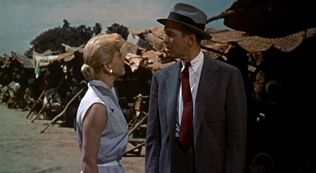 The Man Who Knew Too Much (1956) movie still: medium — An outdoor market setting features rows of woven reed stalls under a bright sky. A man in a…