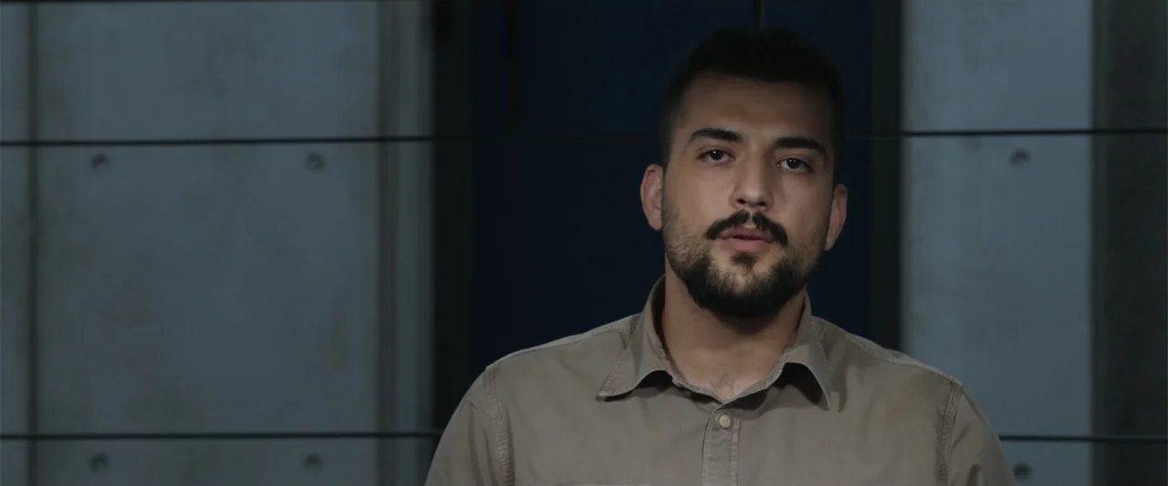 Black Site (2022) movie still: medium — An interior space features concrete wall panels with thin horizontal lines behind a person. A man wearing a…