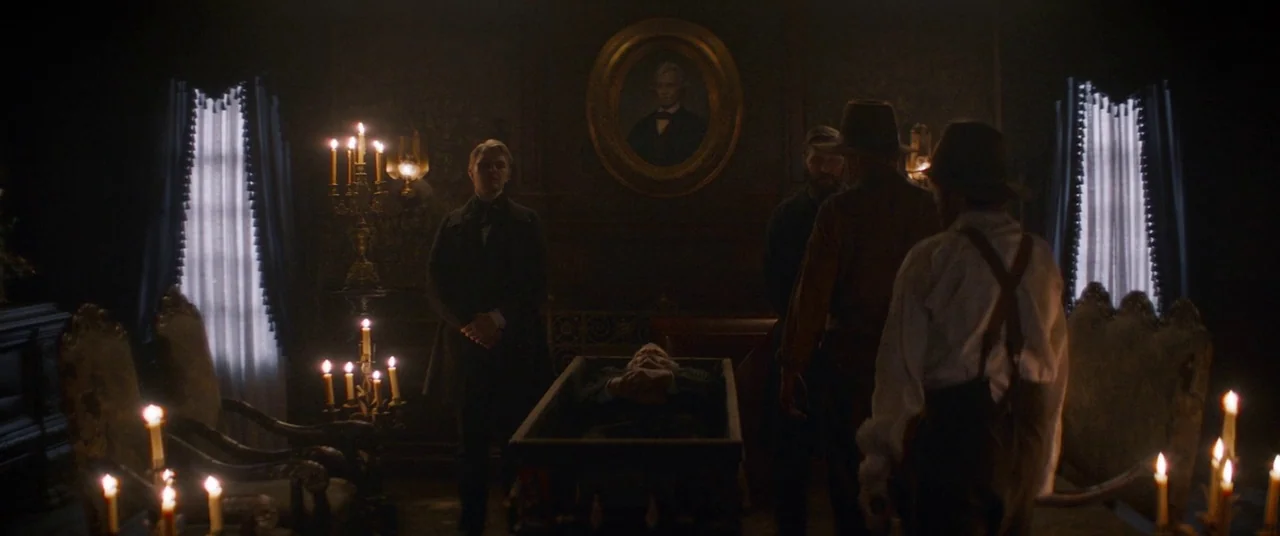 The Sisters Brothers (2018) movie still: wide — A dark, dimly lit parlor room contains a wooden casket placed in the center. Three men in vintage…