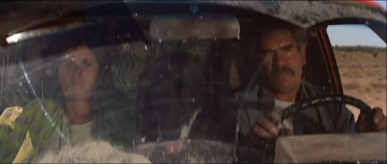 Licence to Kill (1989) movie still: medium — The interior of a vehicle shows two middle-aged adults in the front seats. They appear to be driving…