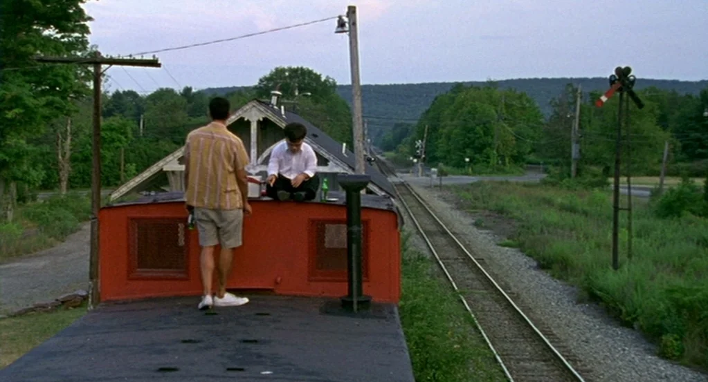 The Station Agent (2003) movie still: wide — A flat-topped red train car sits on tracks surrounded by green foliage and woods. Two men are perched…
