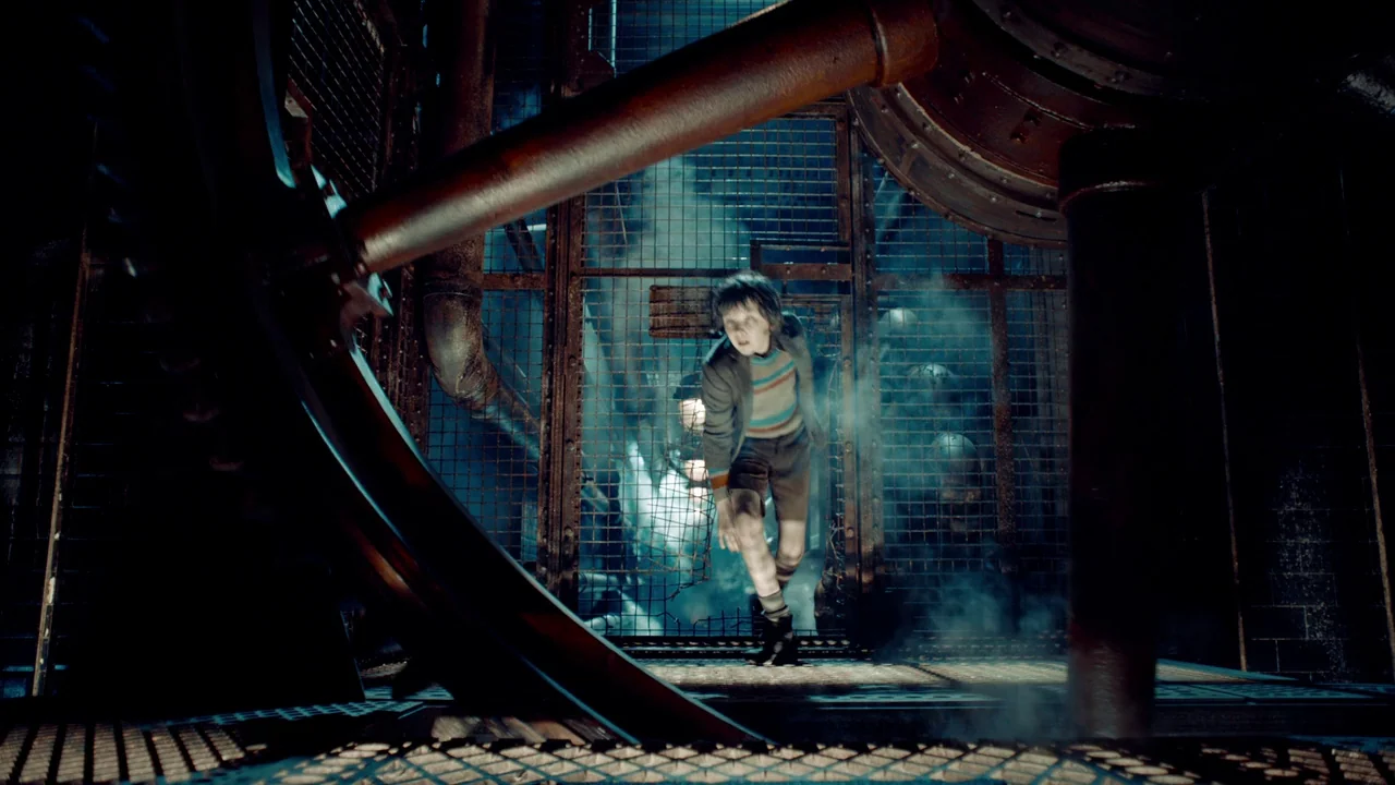 Hugo (2011) movie still: fullbody — A dark industrial setting features large iron gears and a heavy metal safety cage. A young boy in shorts and a…