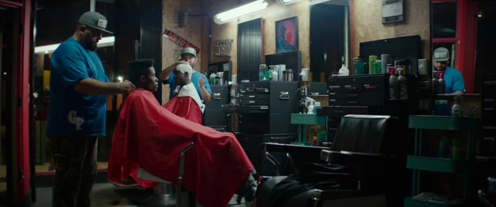 Dope (2015) movie still: medium — A well-lit room with plywood walls, metal equipment cabinets, and multiple barber stations. A barber in a blue…