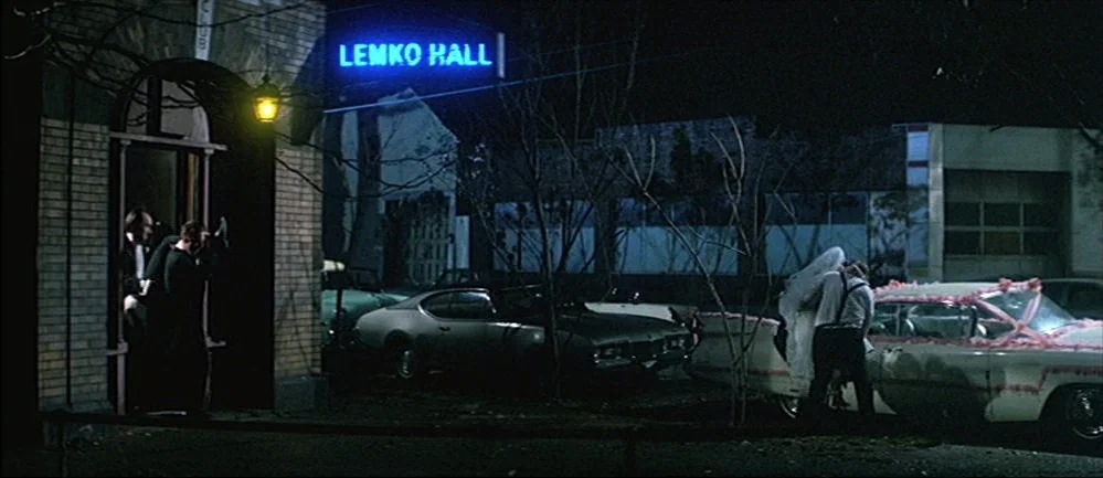 The Deer Hunter (1978) movie still: wide — A brick building entrance at night is illuminated by a blue neon sign. People walk toward vehicles…