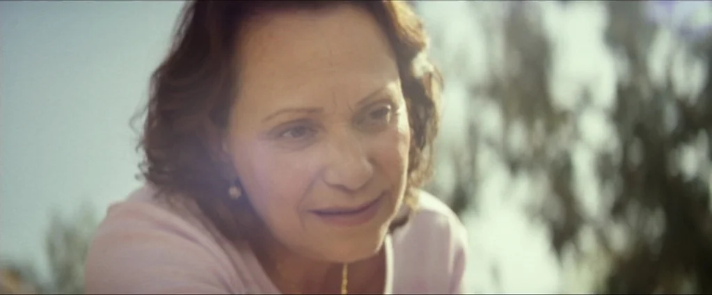 Cake (2014) movie still: closeup — An outdoor area with blurred foliage visible in the background under bright sunlight. An older woman looks down…