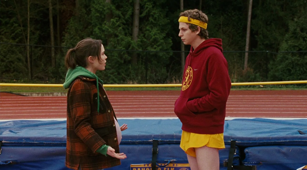 Juno (2007) movie still: fullbody — A high school running track with a high jump mat sits before dense trees. A pregnant girl and a boy in athletic…