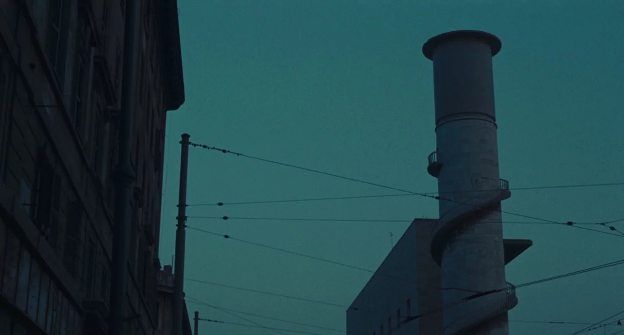 Roma (1972) movie still: establishing — An urban environment features a towering industrial chimney with an external spiral staircase. Overhead…