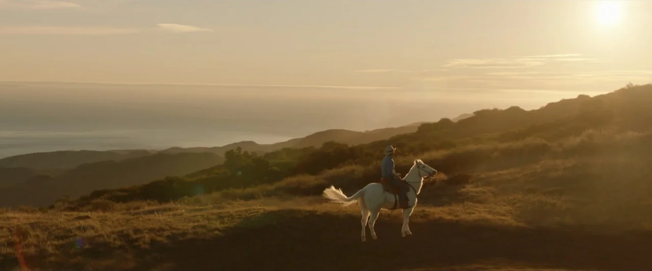 Reagan (2024) movie still: wide — A lone rider on a white horse traverses a grassy coastal hillside at sunset. The ocean is visible in the distant…
