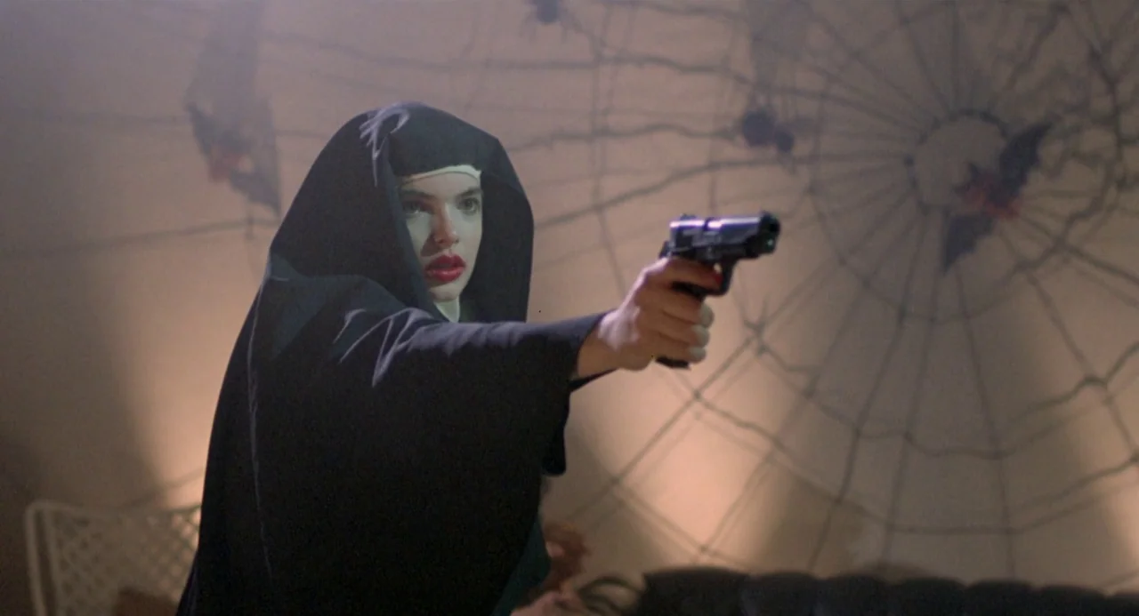 Ms. 45 (1981) movie still: medium — A person dressed as a nun stands before a wall covered in decorative spiderwebs. She aims a black semi-automatic…