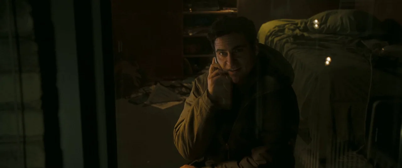 Two Lovers (2008) movie still: medium — A dimly lit room with a made bed in the background and soft focus. A man sits in the foreground using a cell…