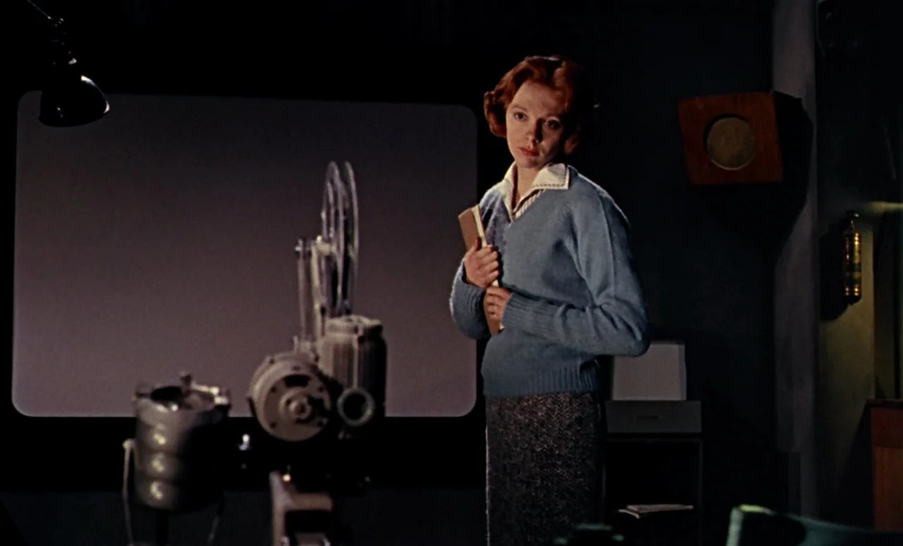 Peeping Tom (1960) movie still: medium — A dark projection room contains a film projector in the foreground and a large screen. A woman stands in…