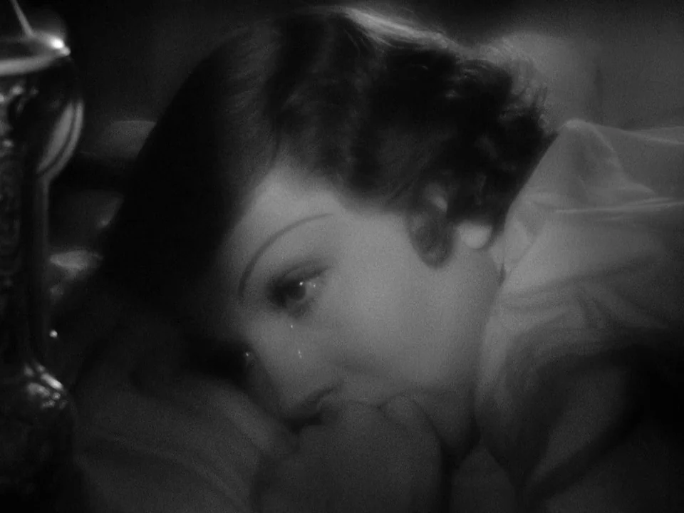 It Happened One Night (1934) movie still: closeup — A dimly lit room provides the backdrop for a woman resting in bed. A person lies on her side,…