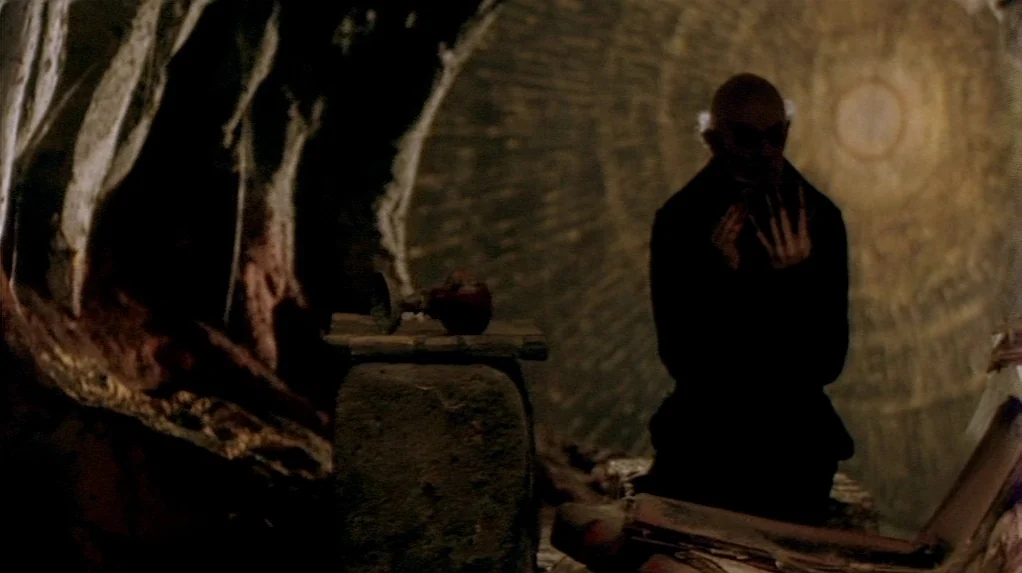 Shadow of the Vampire (2000) movie still: medium — A dark underground stone chamber with textured circular walls and a stone pedestal. A bald man in…