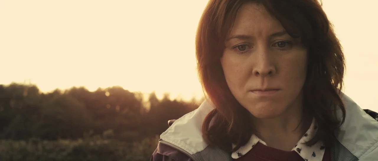 Sightseers (2012) movie still: closeup — An outdoor field at sunset with blurred trees visible in the far background. A woman in a light jacket…