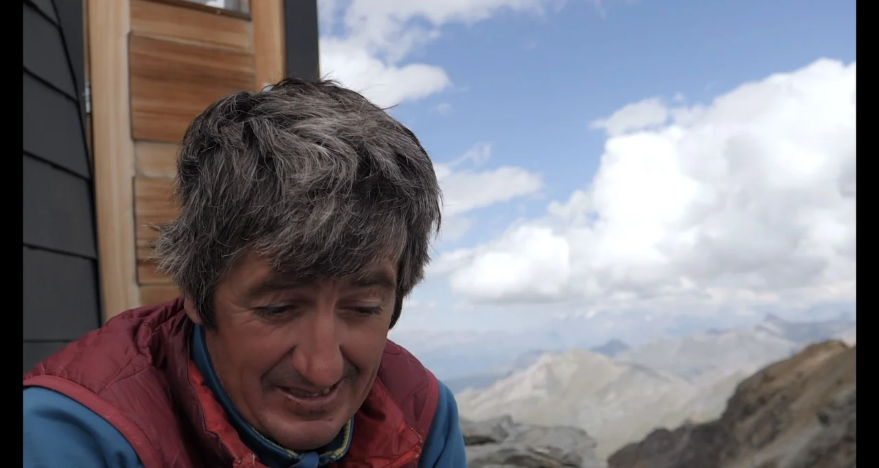 Blutch (2018) movie still: medium — A man stands outdoors near a wooden structure overlooking a vast mountain range. The man wears a quilted vest…
