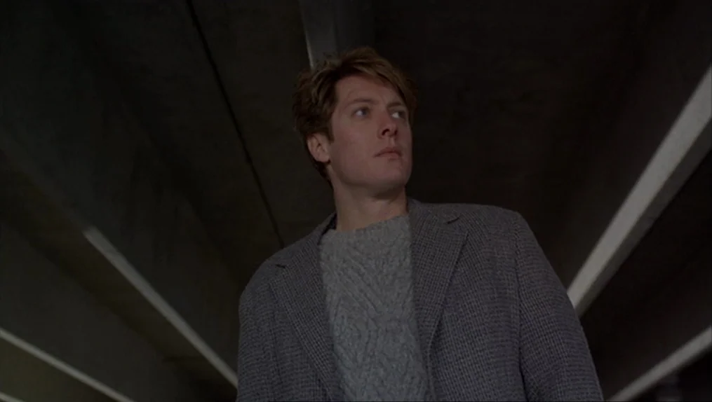 Crash (1996) movie still: medium — A man stands beneath a large grey concrete highway overpass with strong geometric lines. The subject looks…
