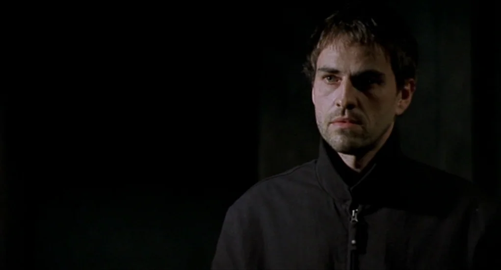 Tiresia (2003) movie still: medium — A dark, empty interior space with no visible background details or architectural features. A man in a dark…
