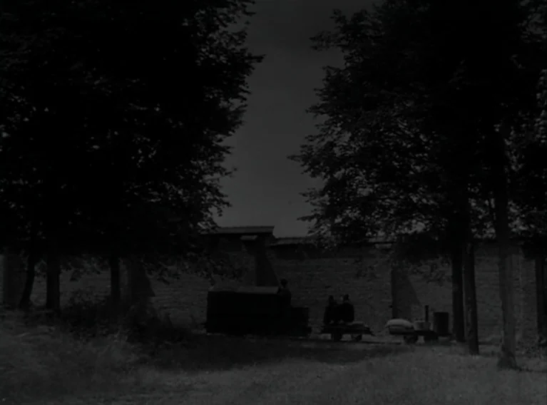 Head Against The Wall (1959) movie still: establishing — A stone wall stands in the background behind a row of dark trees. A small wagon sits on a…