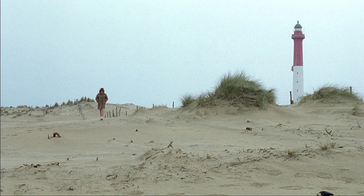 Fat Girl (2001) movie still: wide — A wide expanse of sandy terrain with tufts of grass and wooden fence posts. A woman in a long coat walks toward…