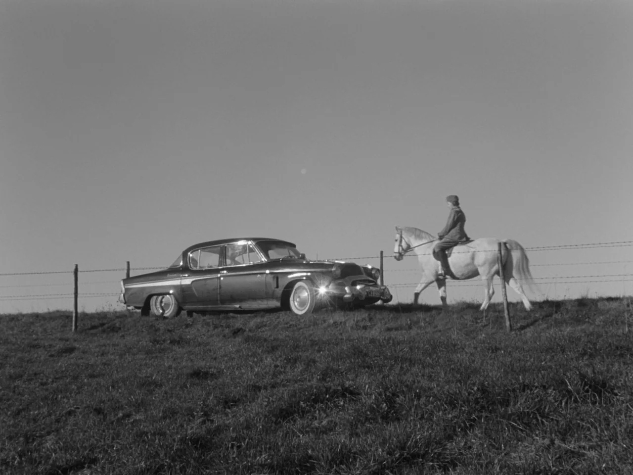 The Baby Carriage (1963) movie still: wide — A grassy hill crest features a barbed wire fence with a parked sedan nearby. A person rides a…