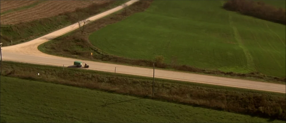 The Straight Story (1999) movie still: establishing — A rural road cuts through vast rolling green pastures and plowed fields. A small lawn mower…