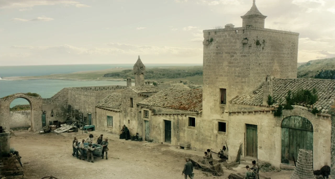 Pinocchio (2022) movie still: wide — A stone village with tiled roofs overlooks the ocean on a clear day. Small groups of people in period clothing…