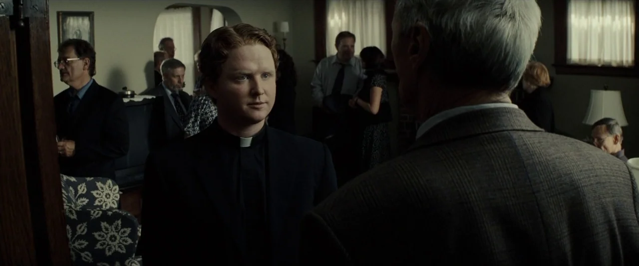 Gran Torino (2008) movie still: medium — A crowded domestic gathering with people socializing in a dim residential interior. A young priest stands…