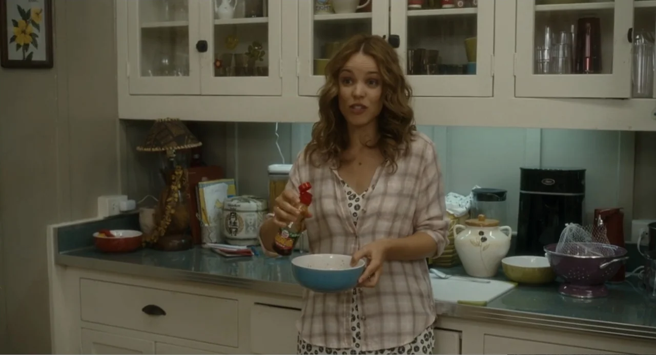 Aloha (2015) movie still: medium — A domestic kitchen with white cabinets features a countertop cluttered with jars and bowls. A woman in a plaid…