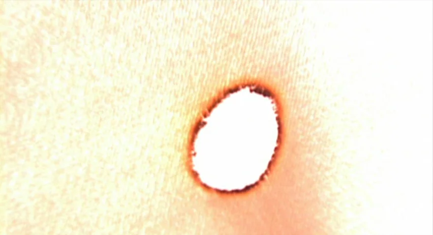 Inland Empire (2006) movie still: closeup — A close-up view shows a small, charred, circular hole in a piece of paper. Light shines through the…